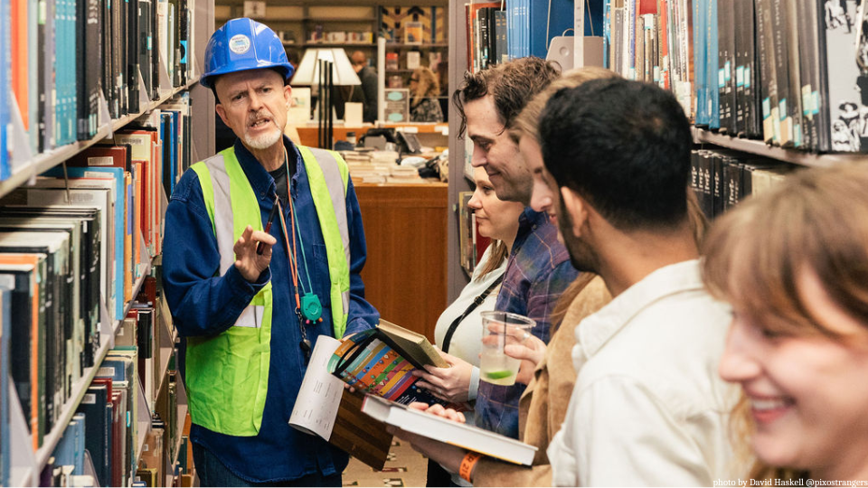 A man in a hard hat in the library stacks instructing guests on a specific process.