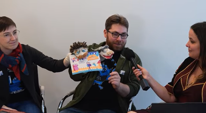 Three people sit in front of a blank wall. One person, holding the microphone, is conducting an interview. The other two figures are holding the German edition of Sock Puppets, each also clad with a sock puppet on their hands.