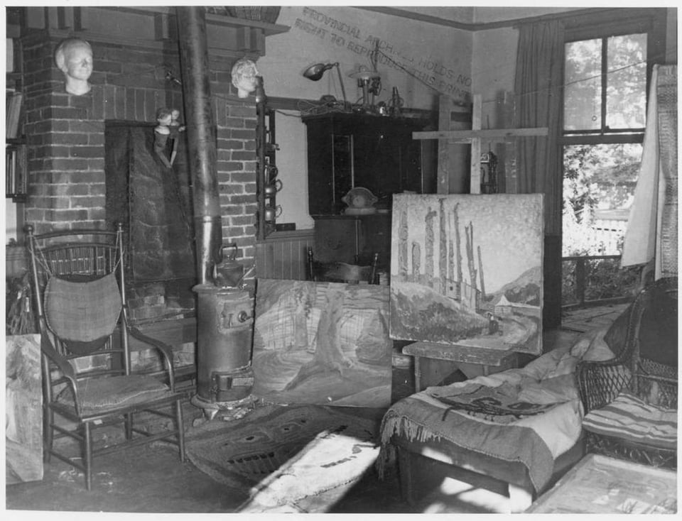 Black and white photo of the interior of a house. There is a small wood stove, a few chairs and a couple of paintings.
