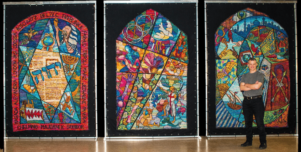 Artist Kirk Dunn (a white man) is wearing a grey t-shirt and a pair of black pants. He is standing in front of a series of knitted tapestries that look like panels of stained glass.
