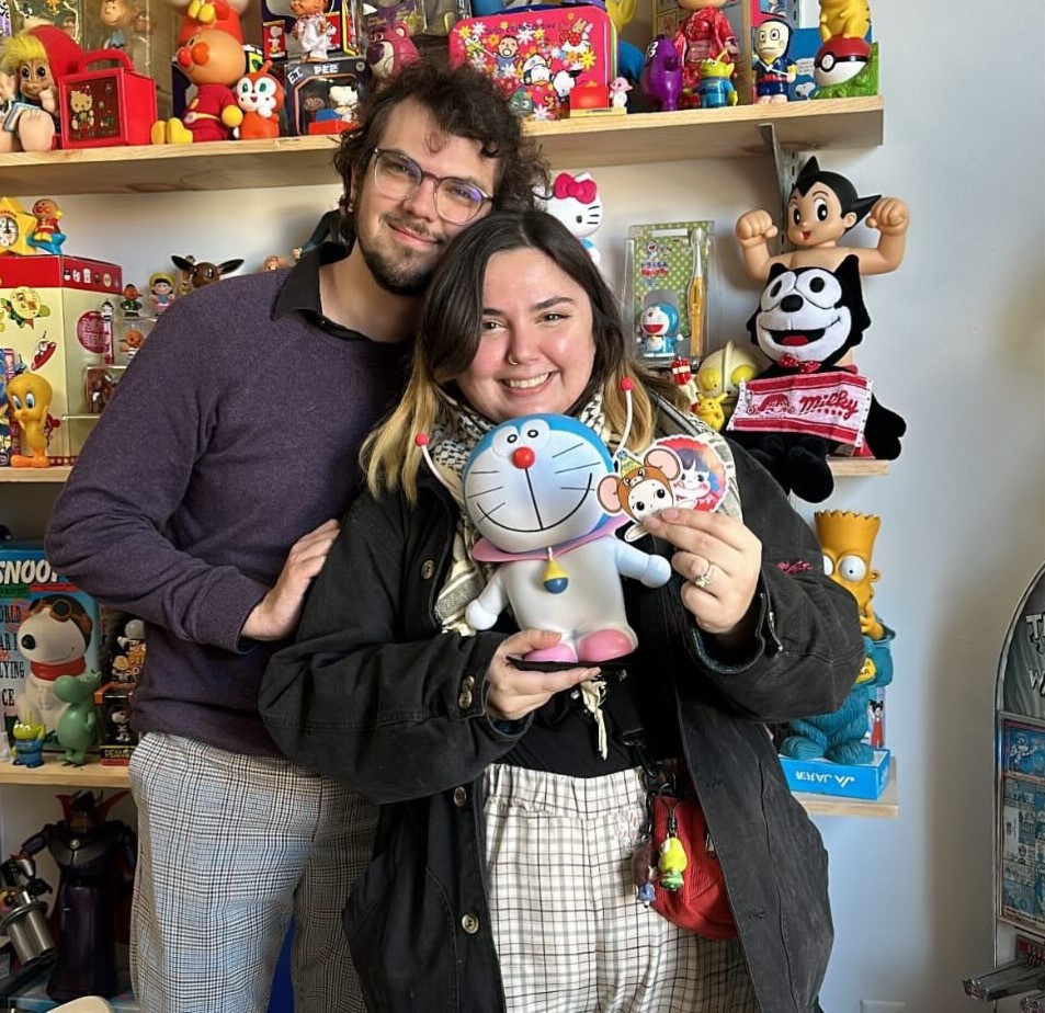 David and Becca stand in MomoToys, an appointment only toy store in LA. Becca is holding a Doremon figure and a pair of cute stickers. They're surrounded by friends like Hello Kitty, Felix the Cat, Snoopy, Astro Boy, and Bart Simpson.