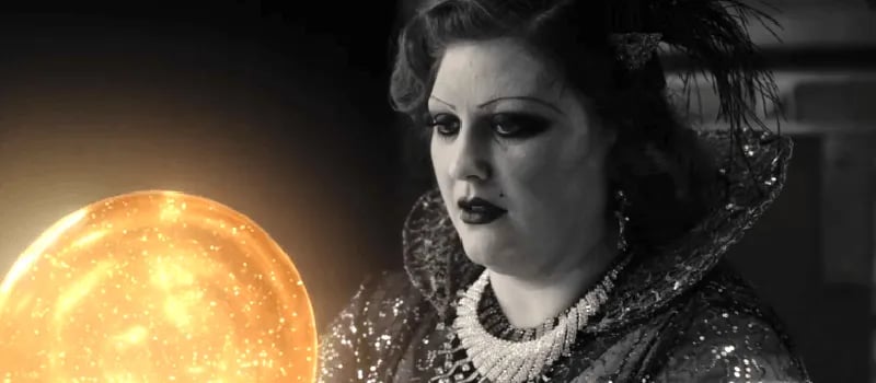 a woman in an ornate dress and necklace, with long penciled eyebrows and deep eyeshadow, looks at the golden orb with an undefinable expression.