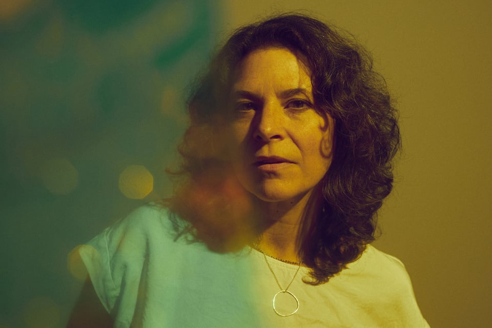 A portrait of a woman with wavy brown hair that reaches her shoulders. She's looking at the camera in muted light, wearing a white t-shirt and a necklace with a circular hoop attached to a chain.