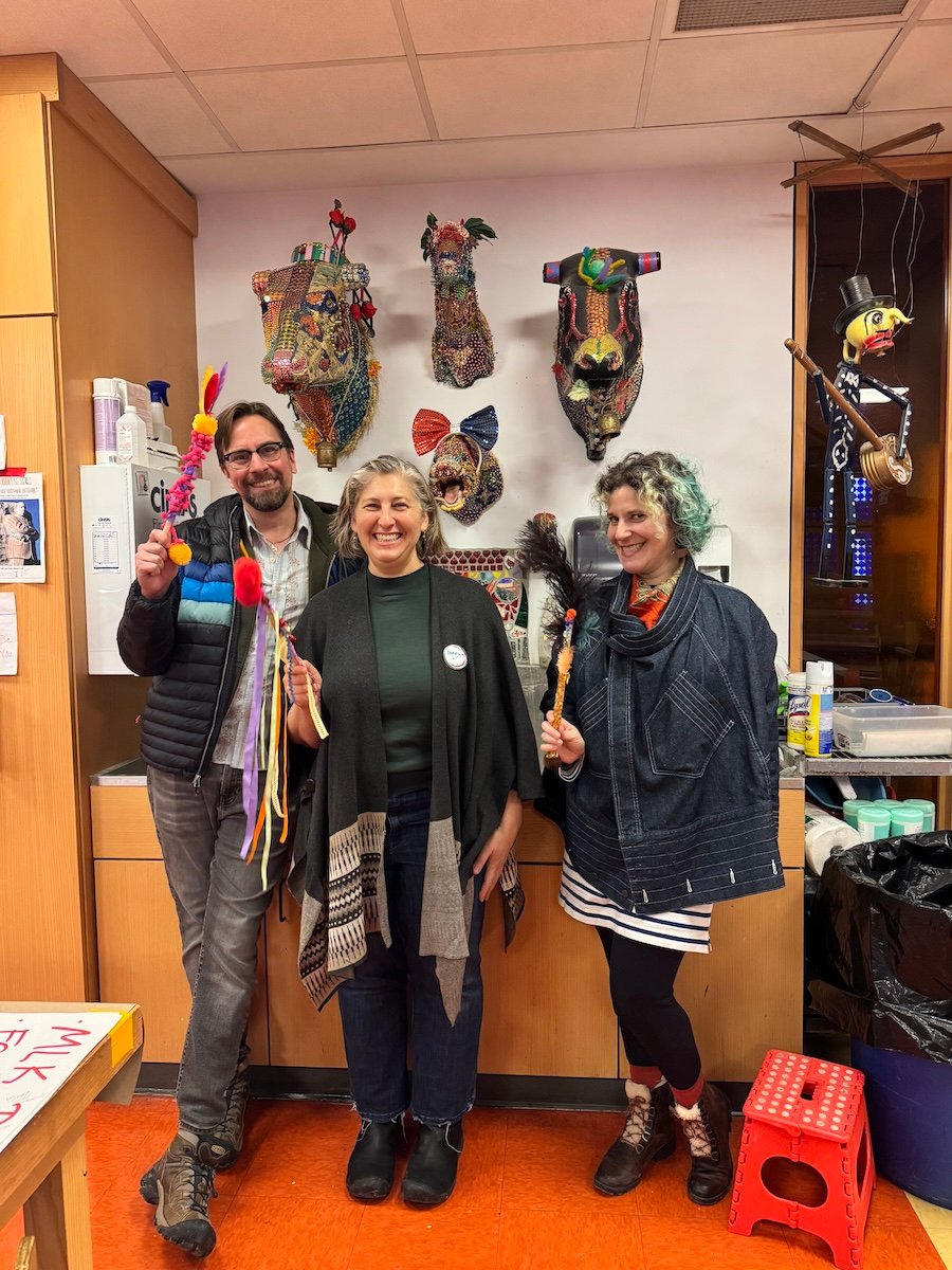 Three people smiling and holding decorated wands, posed in an art studio, with various art pieces adorning the walls, including ornate masks and a puppet.