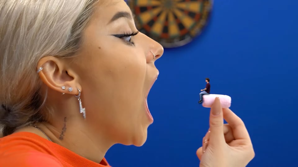 A young blonde woman opens her jaws wide to eat a small pink candy upon which a shrunken man sits.