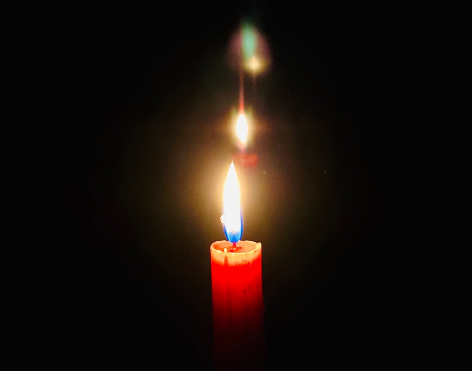 Red taper candle burning in darkness, with echoes of its light reflected behind it.