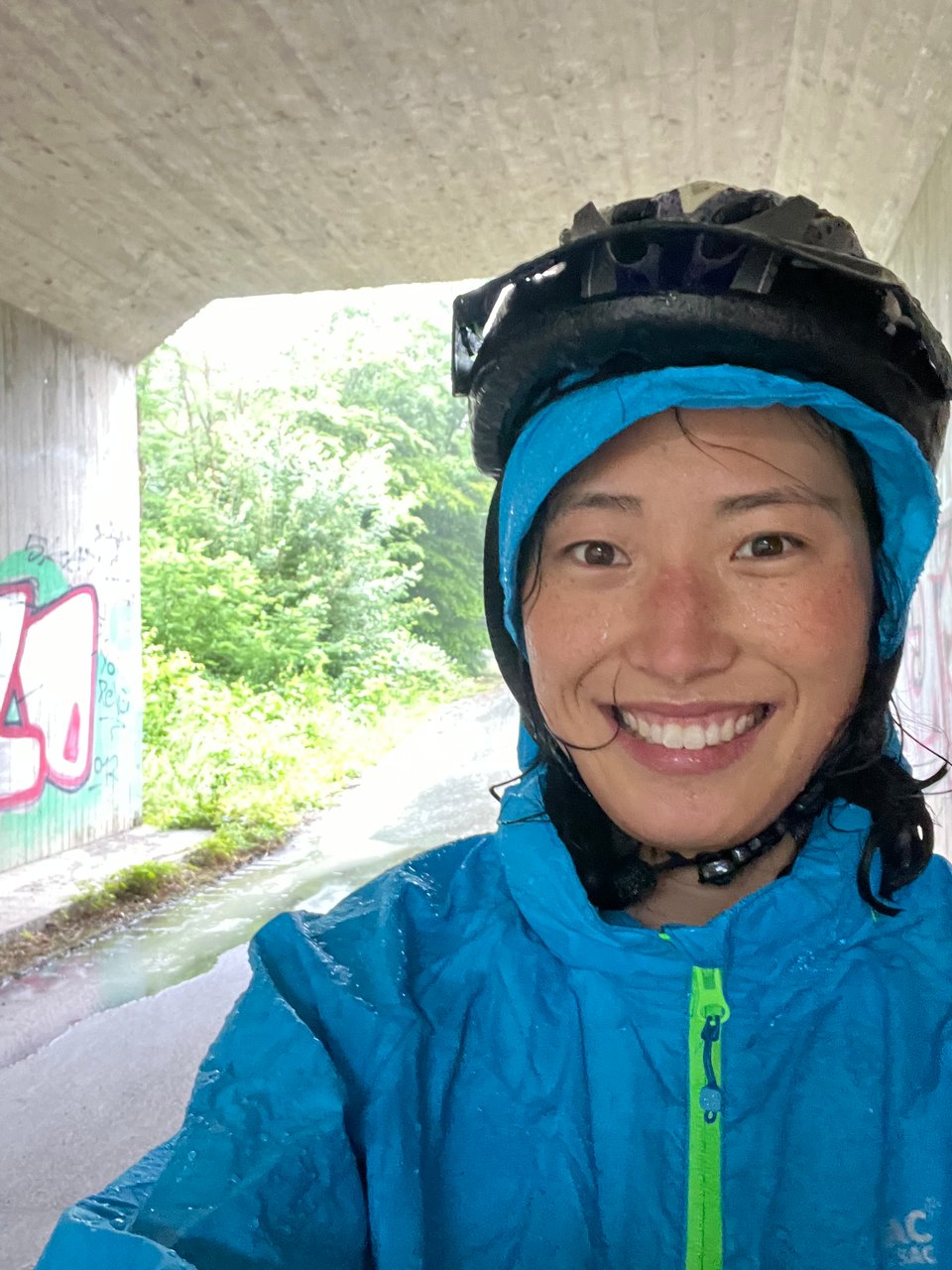 Selfie of Jenny very wet in blue rainjacket and helmet. She's under a tunnel and on the ground behind her is a lot of water flowing past