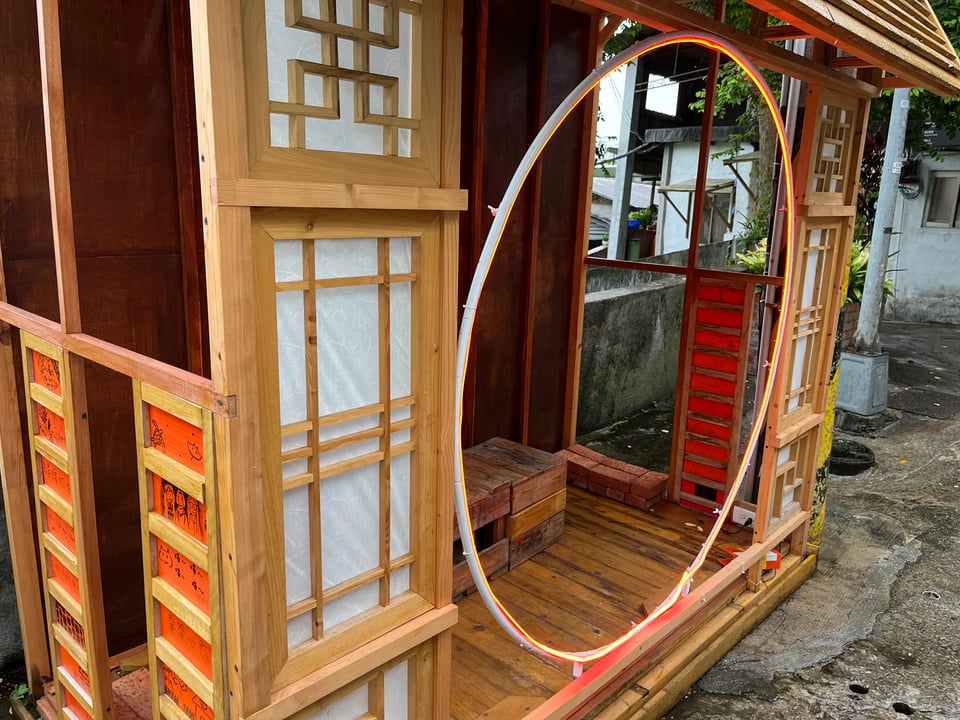 An outdoor pagoda on a stone street. It's made of light wood and red and white paper. It's entrance is framed by an illuminated, circular hoop.