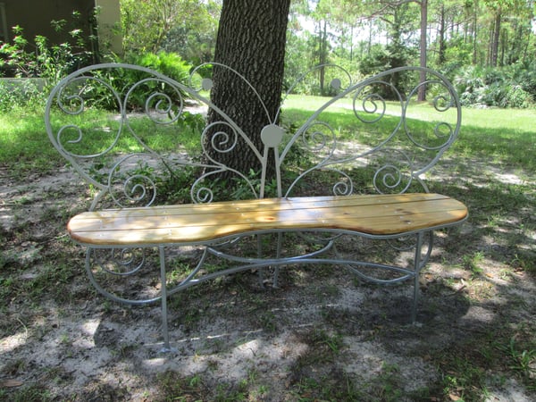 A bench with a wooden seat and a back shaped like a butterfly sits in a shady spot under a tree.