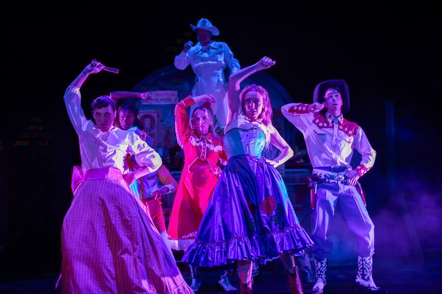A really cool dance sequence in Cowbois that's lit with pink and blue light. The image is of six people dancing in colourful Wild West outfits. From left to right: Emma Pallant, Bridgette Amofah, Sophie Melville, Paul Hunter, Lucy McCormick, Vinnie Heaven. Image credit: Ali Wright.