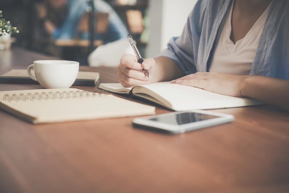 Stock image of a person (face not shown) writing in an open notebook. Beside the notebook on the table is a cup, a phone (screen black), and another open notebook.