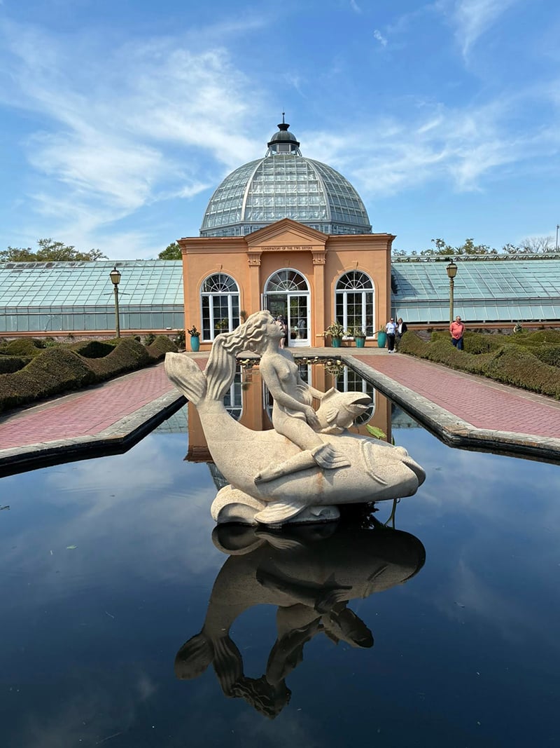 This is a photo from the New Orleans Botanical Garden. There is a building in the background. The foreground is a statue in a pool of water. The statue is of a naked woman riding a large fish. She holds a second, smaller fish to her crotch.