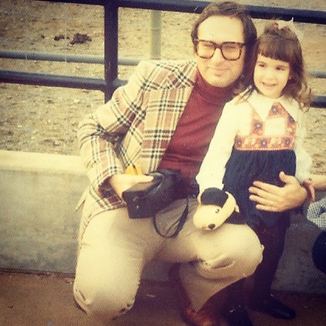 Man in plaid jacket with camera case crouched as he poses with young daughter with dog puppet while visiting the zoo