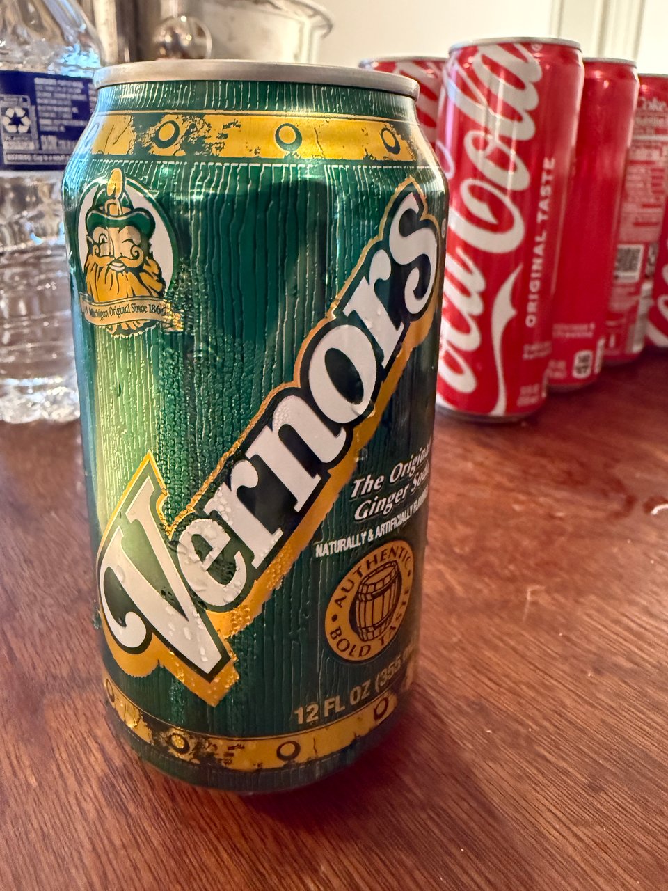 photo of a can of Vernors ginger ale on a wooden table, with bottles of water and cans of Coca-Cola in the background