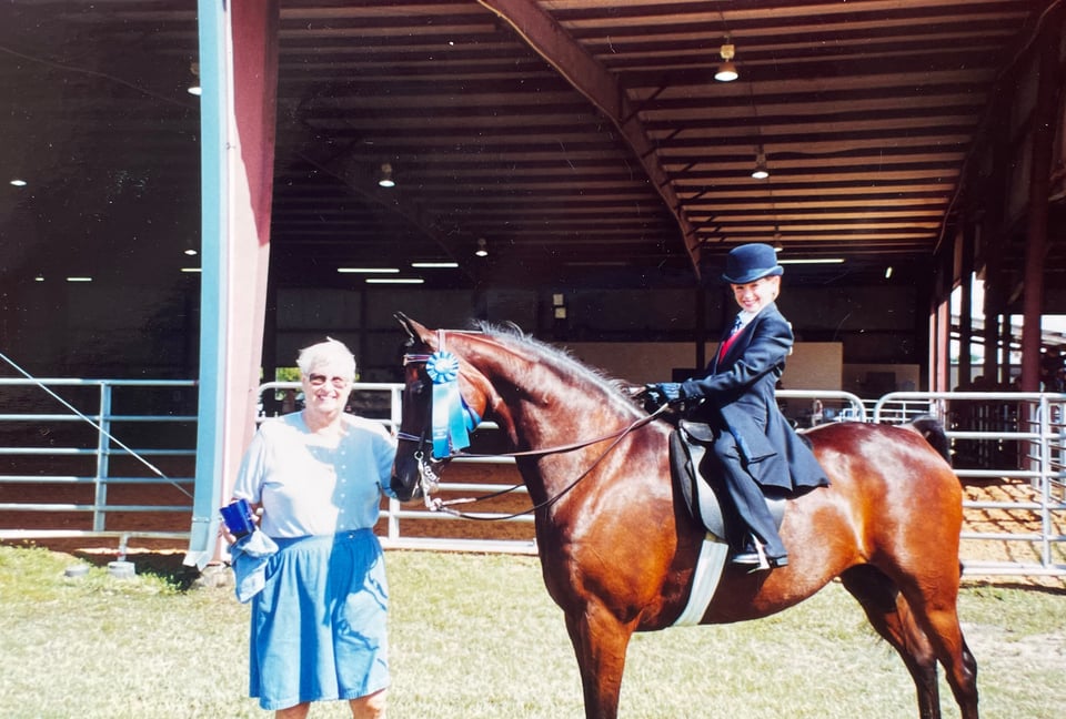 Image of Nana, who is standing by the head of a horse, Tory, that a probably nine-year-old me is riding. I am in a navy suit and hat. There is a blue ribbon hanging from the horse's bridle.