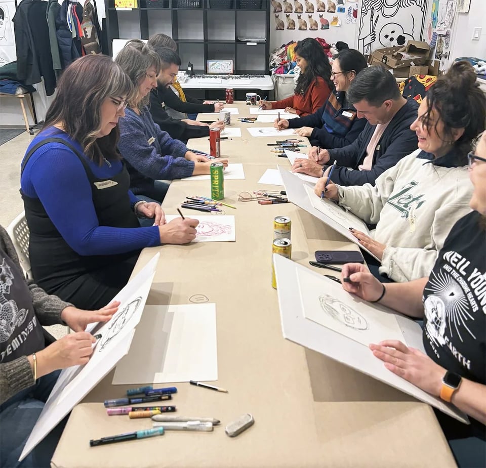 Ten or so people sit at a long table and draw each other