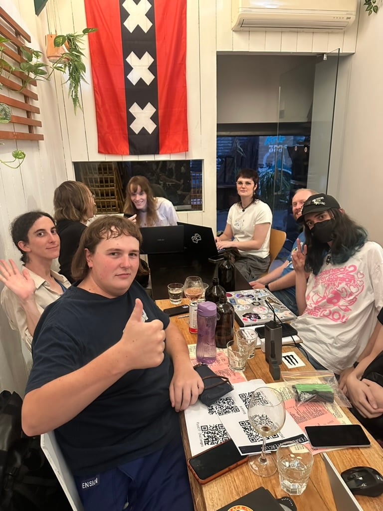A portrait image of several attendees of the Feral Computer Club. Multiple QR codes are on the cable, with laptops open and peace signs all around over a long oak table.