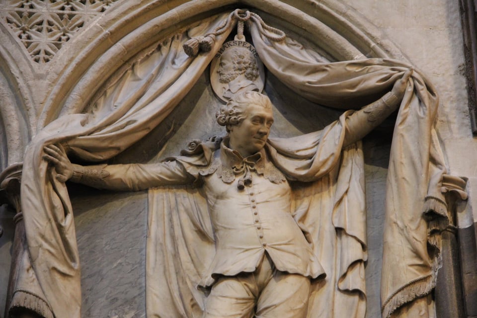 A marble statue of David Garrick, the famous eighteenth century Shakespearean actor, in Westminster Abbey. He's parting a big curtain and wearing a fancy cape, and looking sassy in a big bolo tie and lacey collar.