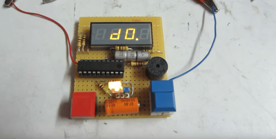 An electronic device, seen from above, sitting on a white table. The electronics are all exposed: Mounted onto a yellow chip board are one blue button, one red button, a microchip, a bright LED, and several resistors and capacitors. It also has an LED number display showing the letter "d" and the number zero.
