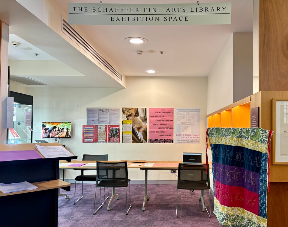 A view of The Just Futures Co-lab Pop-Up at The Schaeffer Fine Arts Library Exhibition Space at the University of Sydney. The image comprises several displayed objects such as a hand-stitched lab banner, tabletop documents, a laptop computer doubling as an interactive hypertext, wall posters, and a screenshot of a video playing on loop on a tv monitor.