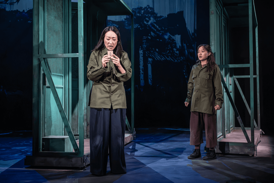 Two women in military-style coats perform on stage, with one holding an object and appearing anxious while the other watches.