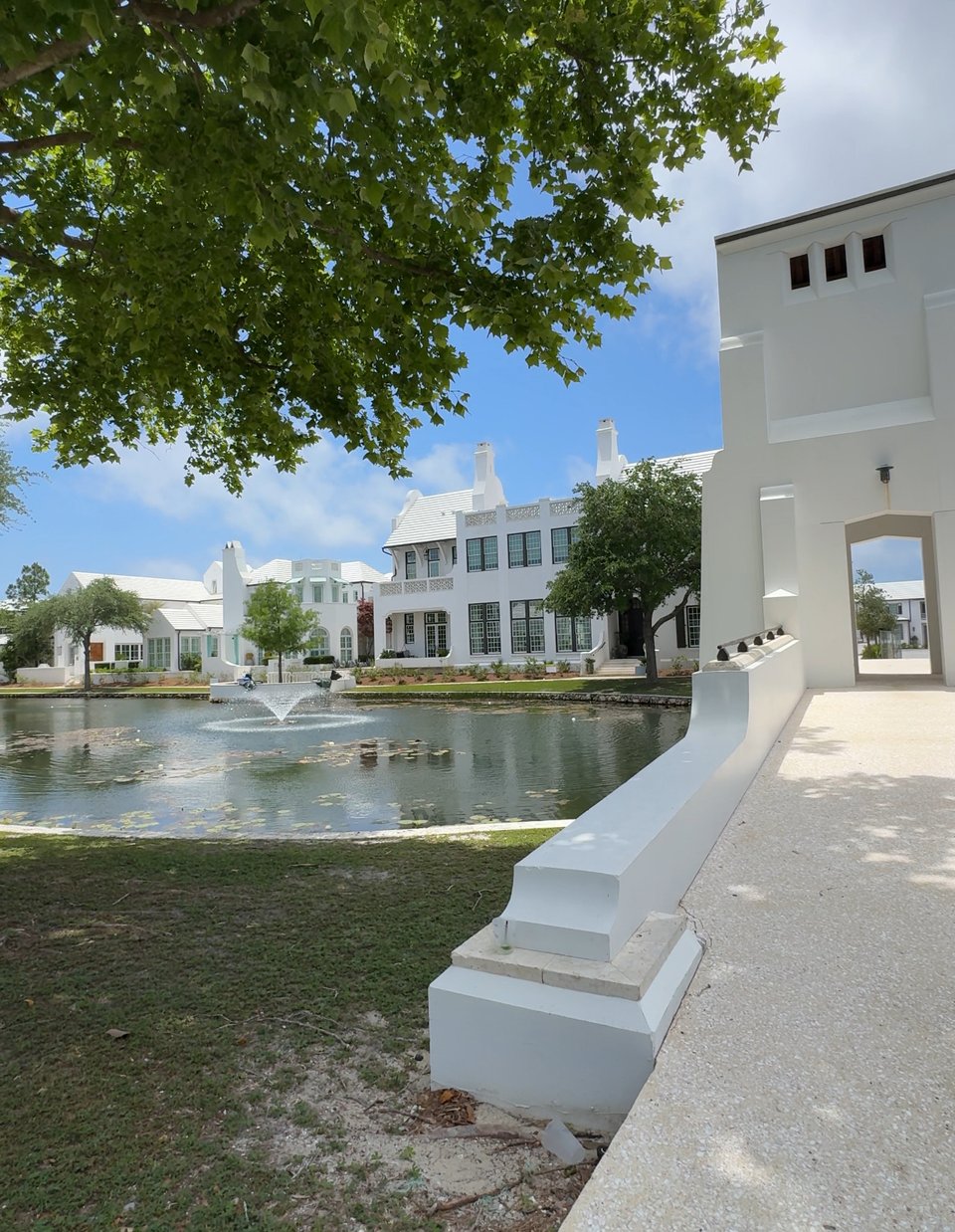 Sunny scene with blue sky and trees. A pond with a fountain surrounded by white homes.
