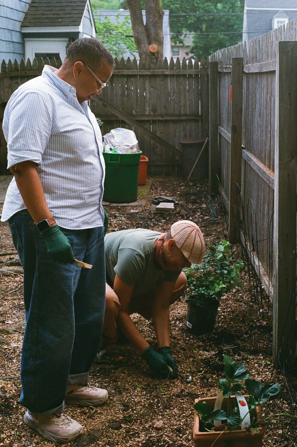 two black women outside with gloves on. one is kneeling with a small shovel, making a hole. the other stands beside. a strawberry plant and blueberry plant are on either side.