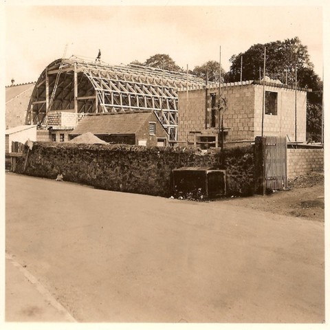 A sepia photo showing the curved wooden roof under construction along with a block built small building next to it. A man is standing high on the curved roof.