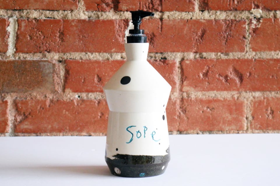 A black and white bottle with a pump dispenser top with 'sope' carved on the side in a child's handwriting. The bottom is black clay, but it shifts with a hard edge slightly spiraled to white clay about two inches up.