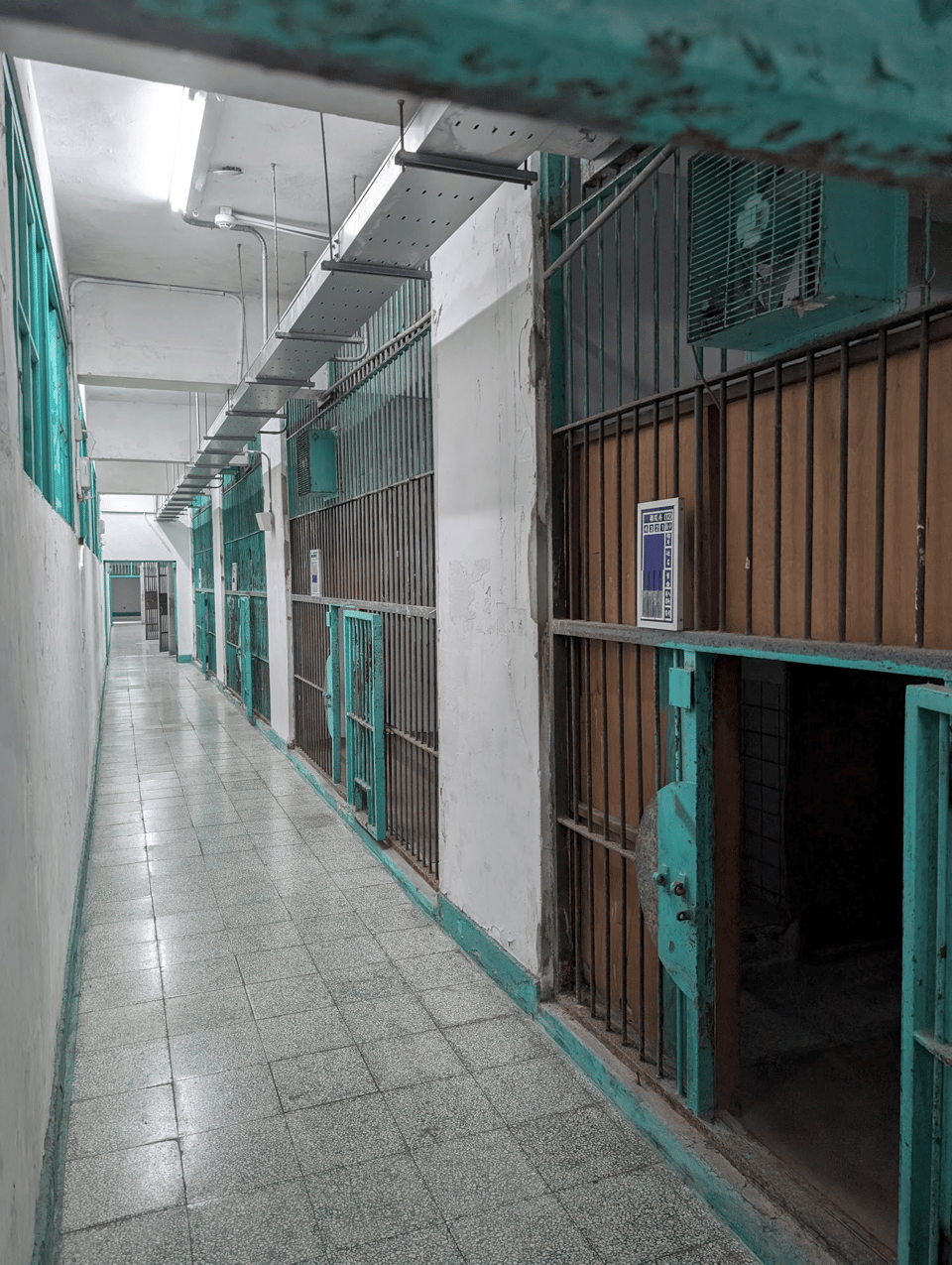 A hallway of prison cells. the paint is aqua green and peeling and rusty-looking. The doors are unusually small, maybe about half the height you'd expect them to be.