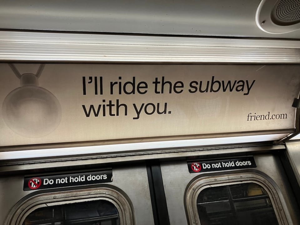 A New York City subway ad with the text "I'll ride the subway with you. friend.com" accompanied by a larger-than-life photo of the Friend surveillance pendant.