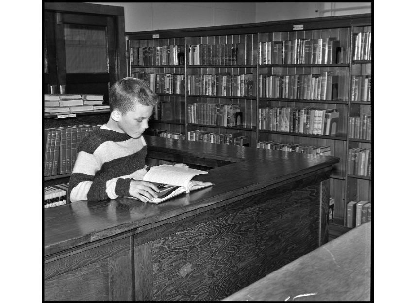 black and white image of a boy in a striped sweater reading a book in a library