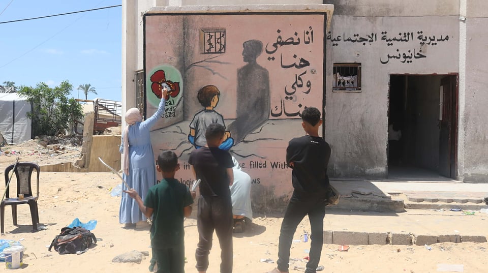 A photo showing an artist painting a mural on the wall of a building in Gaza, illustrating a child sitting on a prison cell floor and looking at their tall shadow cast on the wall, like an adult's shadow. There is a barred window with barbed wire above the child, and the text "أنا نصفى هنا وكلي هناك". Three young kids are photographed from behind, watching her paint.