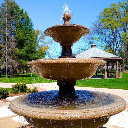a three-tiered concrete fountain burbling merrily with a green lawn, trees, and gazebo in the background against a blue sky