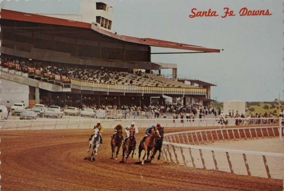 Photo postcard of Santa Fe Downs in the early 1970s. Horses racing around a bend in the track. People scattered throughout the grandstand.