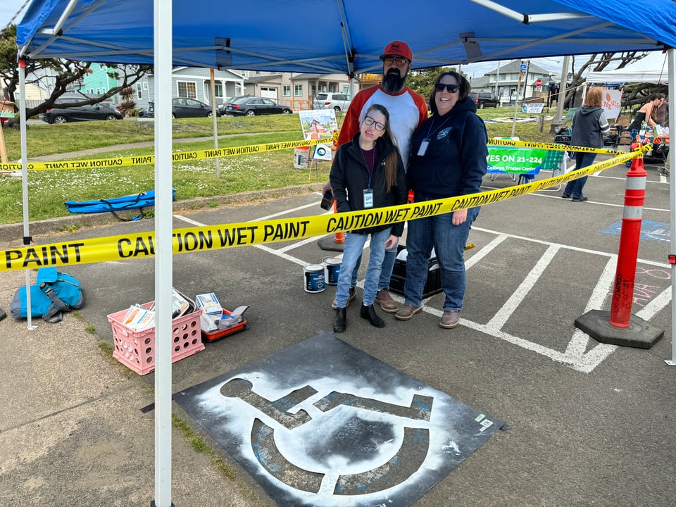 Group of three people, standing in a parking lot with yellow caution tape around a parking space with a paint stencil for an accessible parking space on the ground in front of them.