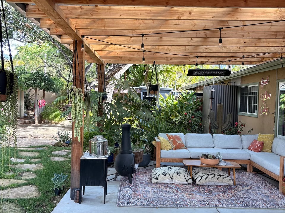The backyard of Andrea's Airbnb. There's a brown, wooden awning over a cream couch, large fluffy floor pillows on top of a multi-colored rug, and twinkling lightbulbs hanging over top. There is also a stone path leading up to more plants, trees, and a chicken coop.