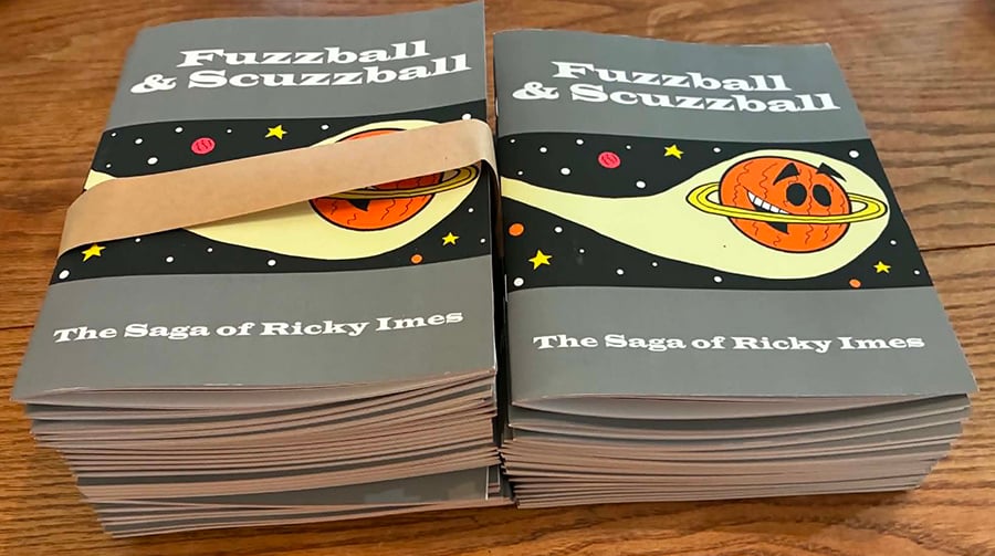 Two stacks of my comic book The Saga of Ricky Imes sit on a wooden table. The cover is mostly gray, with a picture of Ricky Imes in a band across the middle. Ricky is an orange planet who looks like Saturn with eyes and a mouth, who is soaring through space.