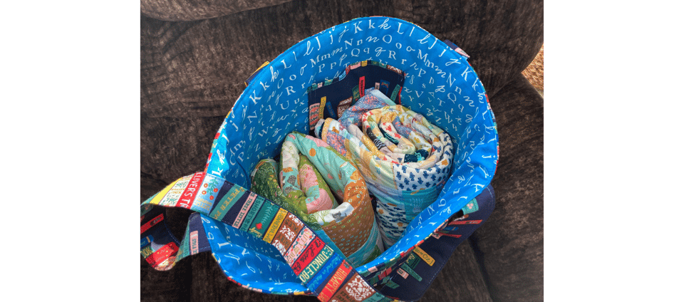 a view looking down into a handmade totebag holding two patchwork quilts rolled up. The tote bag has an interior wiht an alphabet print and the handle is showing, displaying book covers.