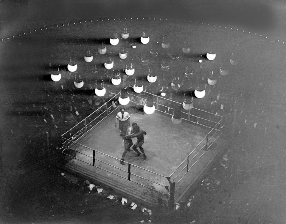 An antique photograph of a boxing match, let by a matrix of early artificial lighting.