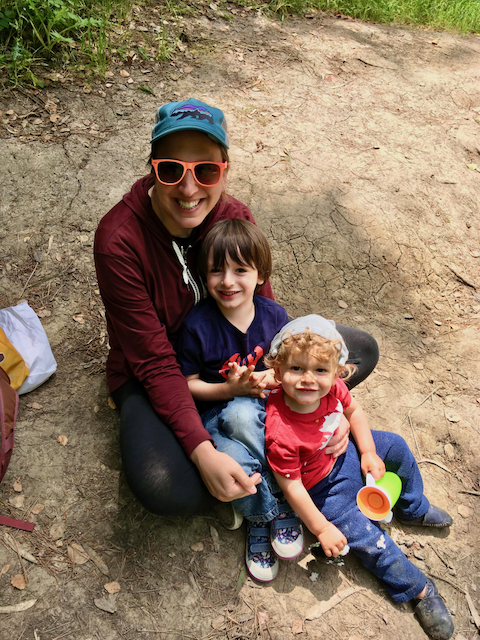 A cute mom wearing sunglasses and outdoor clothing sits on stony ground with two adorable children, one school age brunette, one a curly-haired blond toddler.