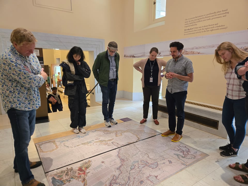 A group of people in a library exhibition space, looking down at a large map displayed on the floor