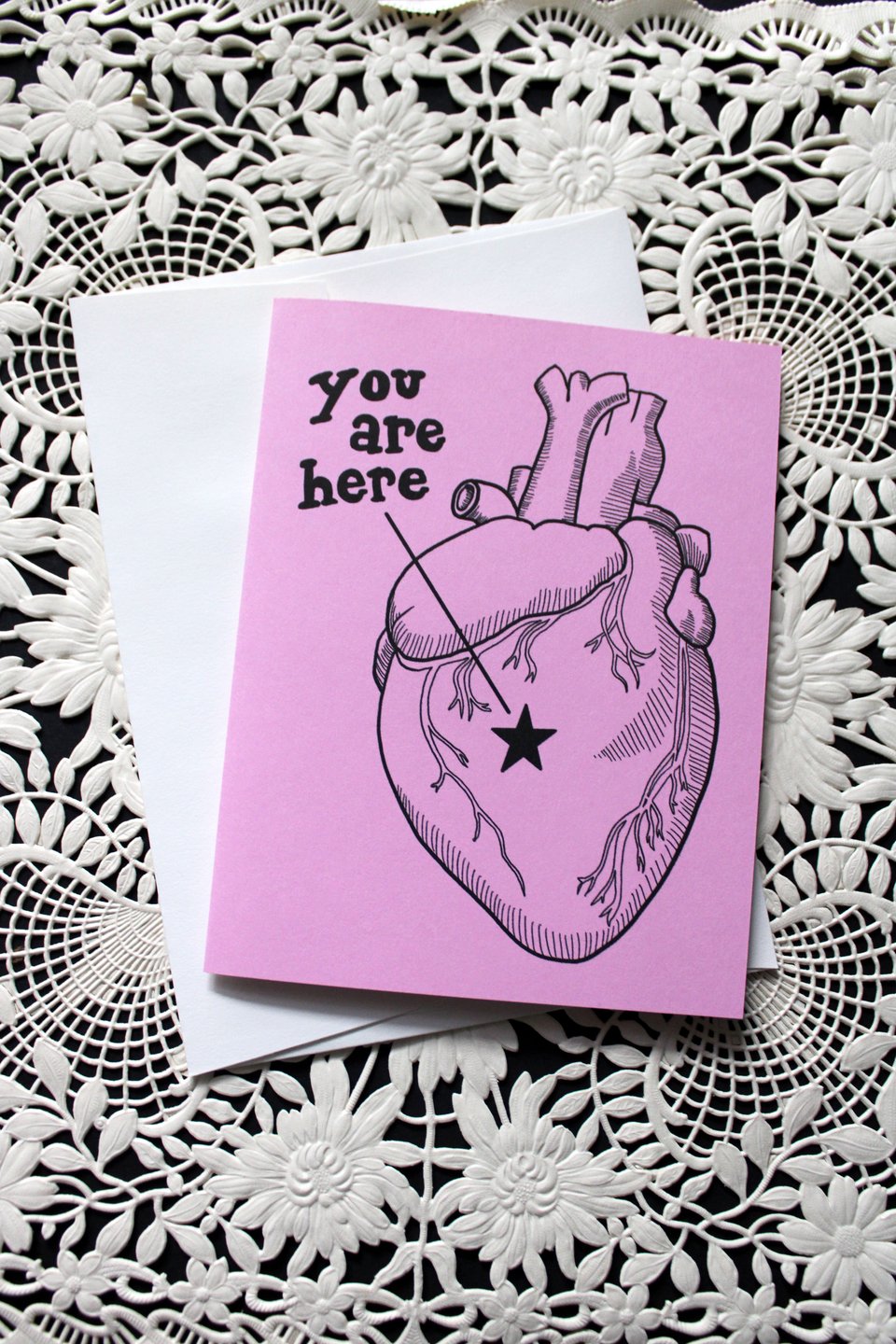 a pink valentine sitting on an ornate paper doily. it’s a line art drawing of an anatomical heart with a star on it and a line running from it that says, “you are here.”