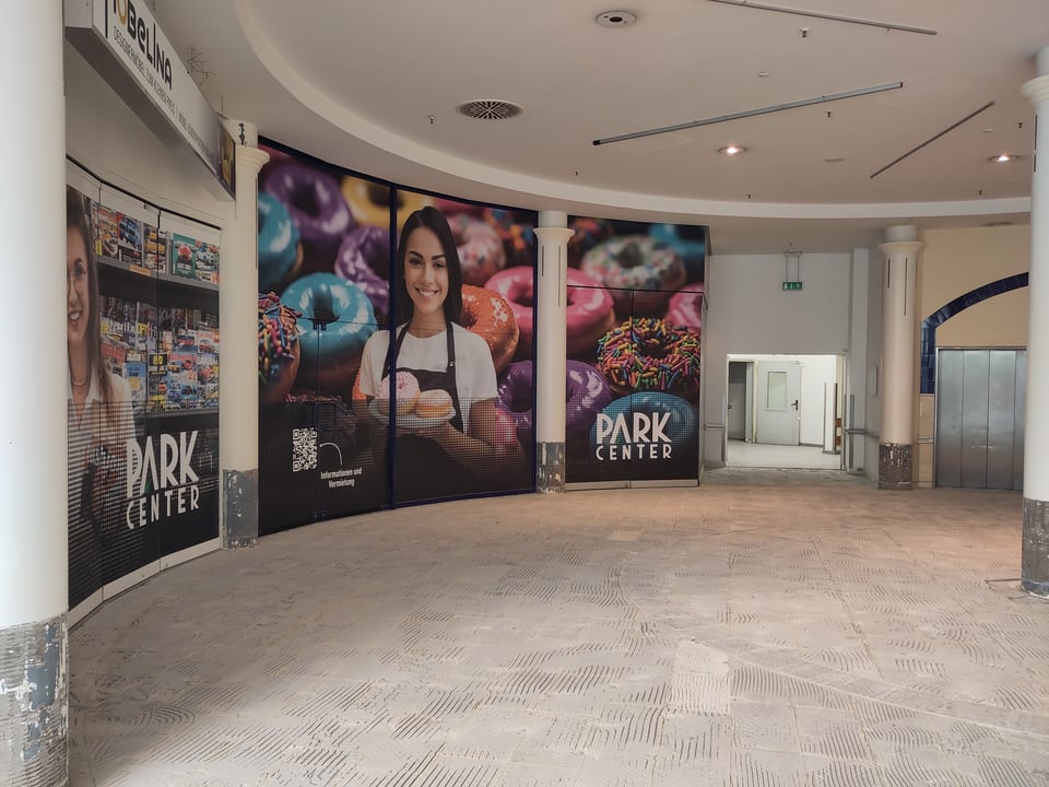 Ausschnitt eines runden, leeren Raums, mit einer geöffneten Tür am fernen Ende, die in einen ebenfalls leeren Gang führt. Im Bogen zu linker Hand generische Werbung den Verkauf von Backwaren (es gibt kein entsprechendes Geschäft im Center), ganz rechts die geschlossene Tür zu einem Fahrstuhl. Der Boden ist entfließt und aufgeraut.