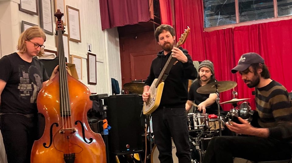 Four musicians, one on upright bass, one on electric bass, one on drums, and one on guitar, performing.