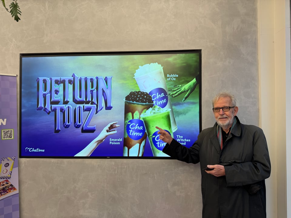 A large monitor shows green and white "cha time" bubble teas with the words "Return to Oz" on it. A man stands next to the monitor and points.