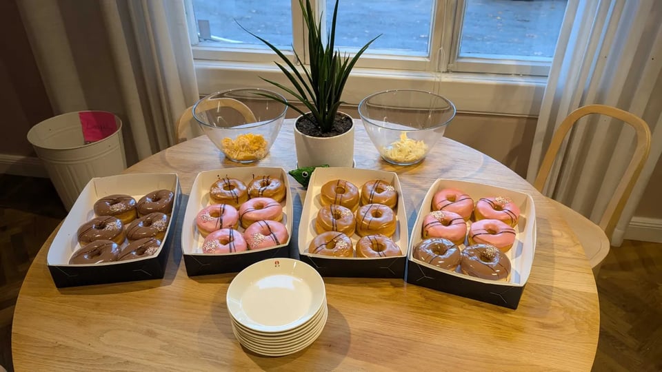 Four boxes of donuts (six in each) on a round kitchen table. Donuts have pink, dark brown and light brown frosting and chocolate stripes on top.