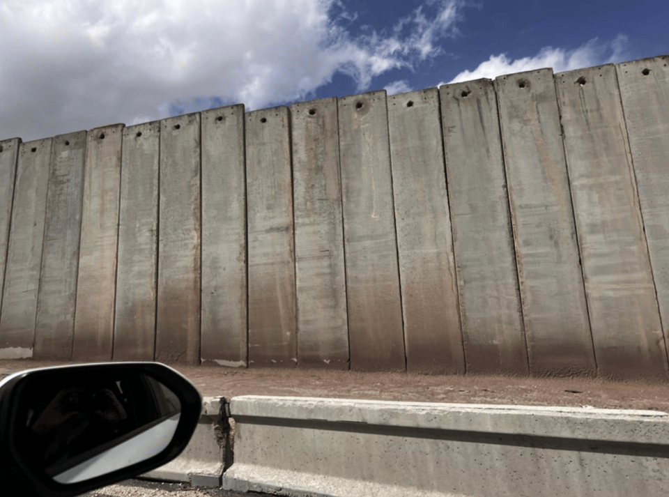 A car's sideview mirror, and a tall concrete wall against a blue sky with white clouds