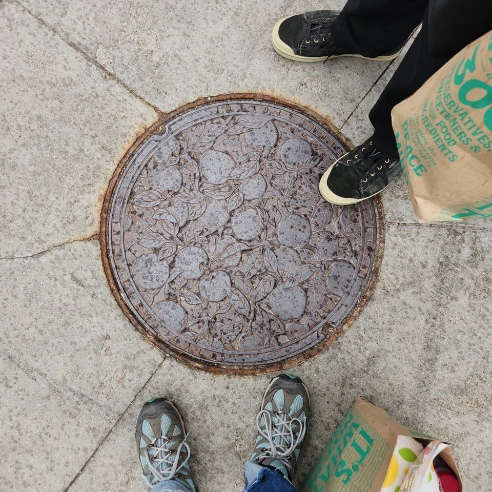 Top-down view of a manhole cover in downtown Minneapolis. The cover shows apples and foliage. Two people's shoes are visible.