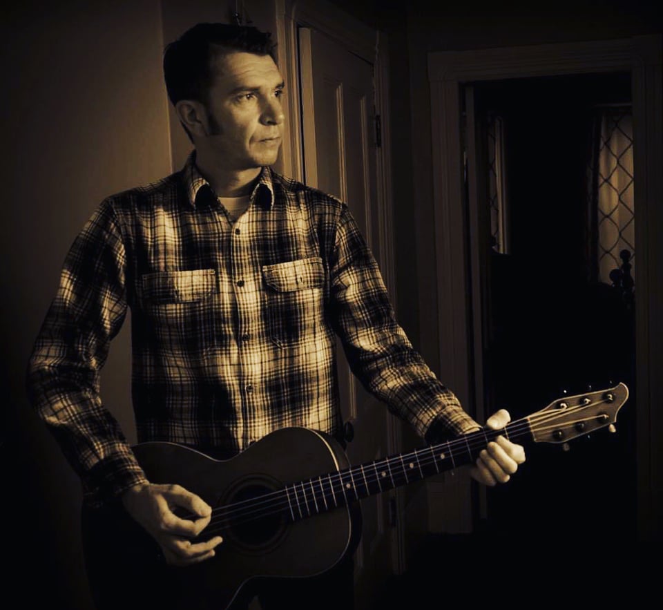 A man with long sidburns wearing a crisp flannel shirt and playing an acoustic guitar looks to his left in a sepia-toned photo.